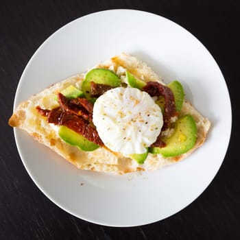 Poached egg on a toasted bun with avocados and dried tomatoes on white plate.