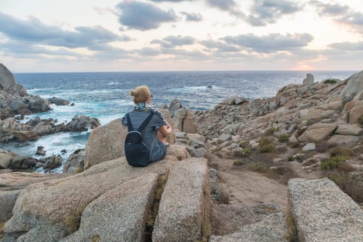 Solitary young female traveler watches a beautiful sunset on spectacular rocks of Capo Testa, Sardinia, Italy, a popular summer traveling destination in Europe.