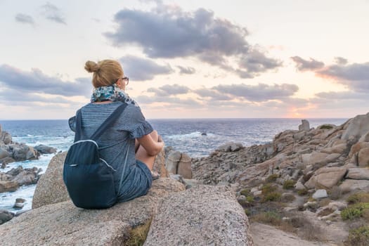 Solitary young female traveler watches a beautiful sunset on spectacular rocks of Capo Testa, Sardinia, Italy, a popular summer traveling destination in Europe.