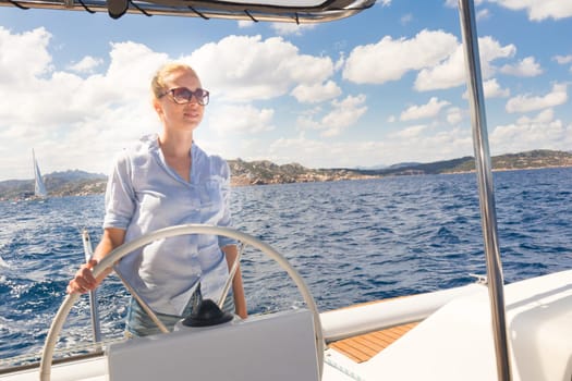 Attractive blond female skipper navigating the fancy catamaran sailboat on sunny summer day on calm blue sea water. Luxury summer adventure, active nautical vacation.