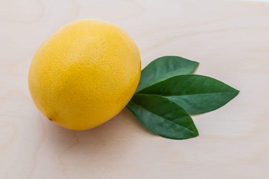 close-up of a yellow bright lemon with three green leaves on a chopping board of light wood.