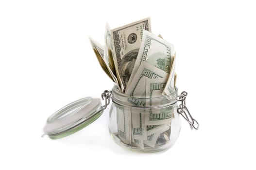 A glass jar filled with dollars among a hundred dollar bills on a white background.