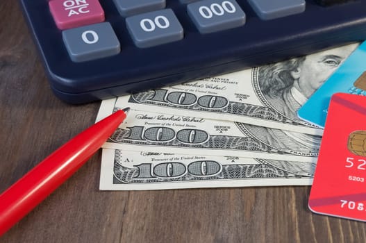 Banknotes for one hundred dollars, credit cards, a calculator and a pen on a wooden table.
