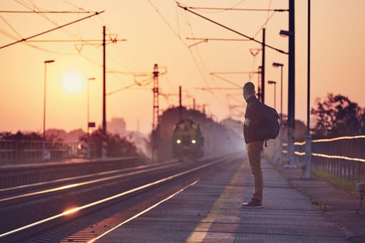 Alone man with backpack waiting at railroad station platform at sunrise.