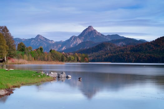 Long exposure of a landscape view of the Weissensee in Bavaria surrounded by autumn deciduous trees. In the background, the summit of the pillar rises into the cloudy sky.