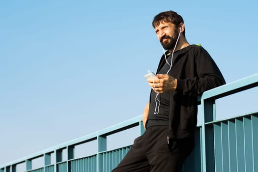 The bearded man is using phone with earphones for listening music outdoors.