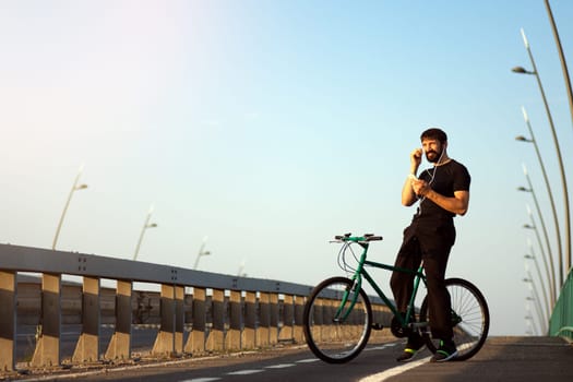 The smiling cyclist is using phone with earphones for listening music outdoors on the bridge.