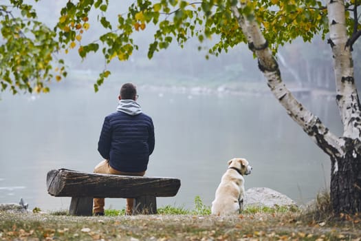 Pensive man with dog is contemplation on wooden bench near pond in autumn nature.