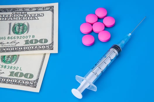 Syringe, dollars and pink pills on a blue background.