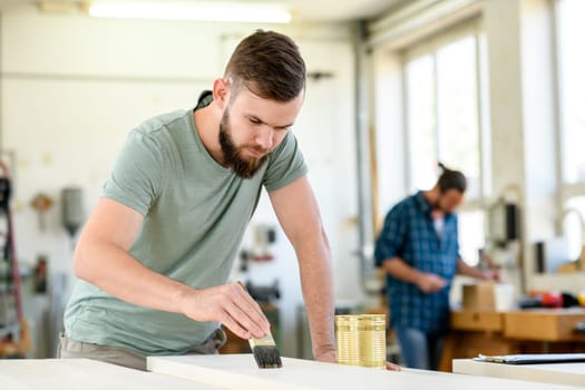 young worker with workpiece in a carpenter's workshop painting wood with brush