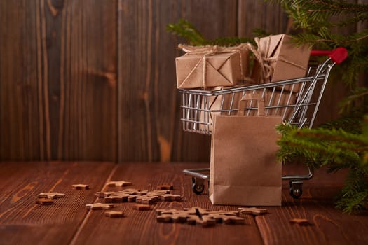Shopping trolley with gift boxes and fir tree branches on a brown wooden background. Christmas and New Year shopping.