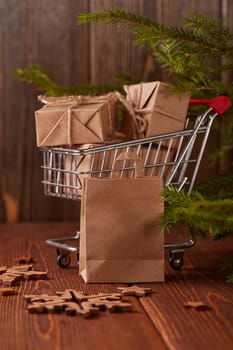 Shopping trolley with gift boxes and fir tree branches on a brown wooden background. Christmas and New Year shopping.
