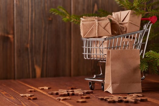 Shopping trolley with gift boxes and fir tree branches on a brown wooden background. Christmas and New Year shopping.