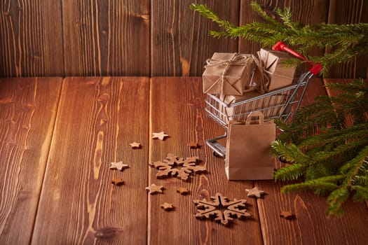 Shopping trolley with gift boxes and fir tree branches on a brown wooden background. Christmas and New Year shopping.