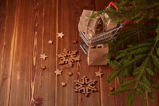 Shopping trolley with gift boxes and fir tree branches on a brown wooden background. Christmas and New Year shopping.