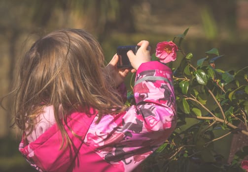 Cute little caucasian girl taking photos of flowers in the park in spring