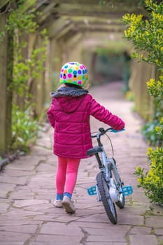 Young little girl pushing her bicycle on the stony retro garden pathway in a public park