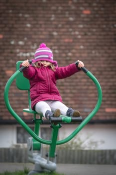 Little Caucasian girl exercising on the outdoor training machine on a chilly autumn day
