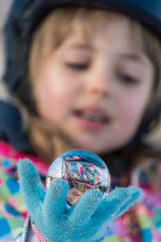 Winter portrait of a little girl wearing ski helmet and holding small glass ball with her own reflection