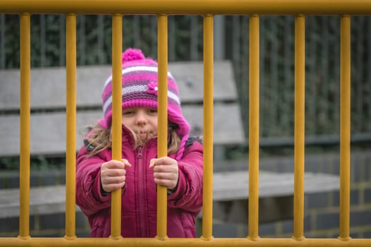 Little cute Caucasian girl dressed in pink hat and jacket standing behind metal gates leading to the playground