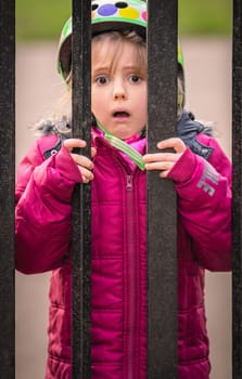 Little cute Caucasian girl dressed in pink jacket standing behind metal gates leading to the playground
