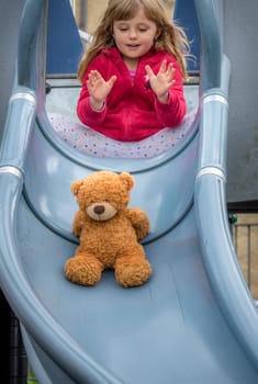 Adorable Caucasian girl pushing teddy bear from a top of a slide while playing on an outdoor playground