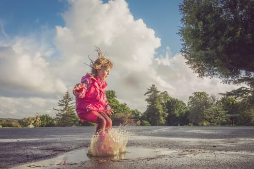 Little Caucasian girl jumping into the puddle after rain in english countryside