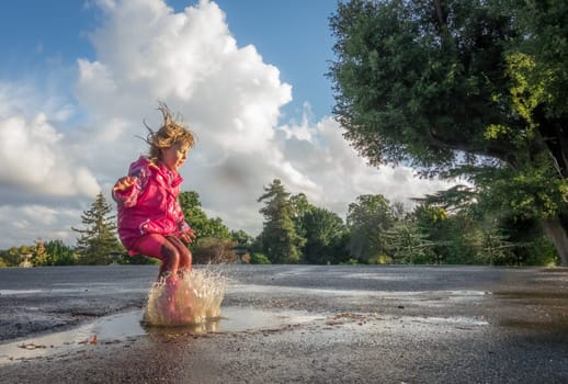 Little Caucasian girl jumping into the puddle after rain in english countryside