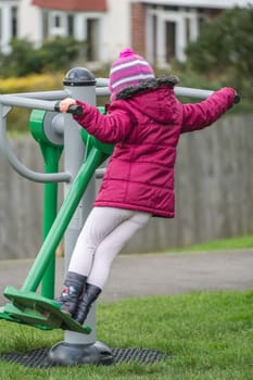 Little Caucasian girl exercising on the outdoor training machine on a chilly autumn day