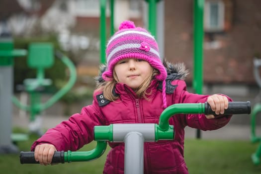Little Caucasian girl exercising on the outdoor training machine on a chilly autumn day
