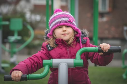 Little Caucasian girl exercising on the outdoor training machine on a chilly autumn day