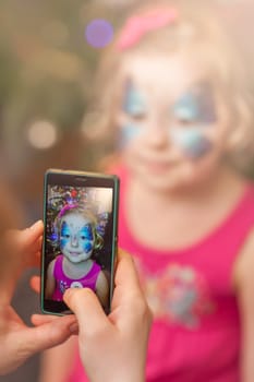 Mother taking picture on a smarphone mobile phone of her little daughter girl having her face painted during party