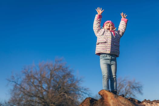 Cute little girl shouting from joy while standing on the top of large cut tree log in the park in spring