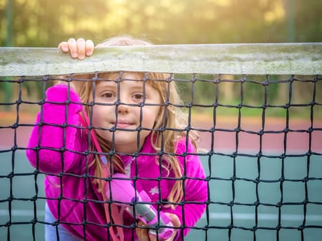 Cute young Caucasian girl dressed in pink jumper and holding toy camera looking through tennis net