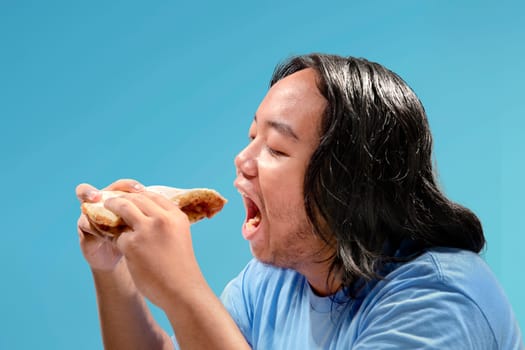 Asian fat man eating slice of pizza fast food over colored background