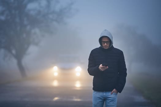 Young man walking on road and using phone. Car approaching in thick fog.