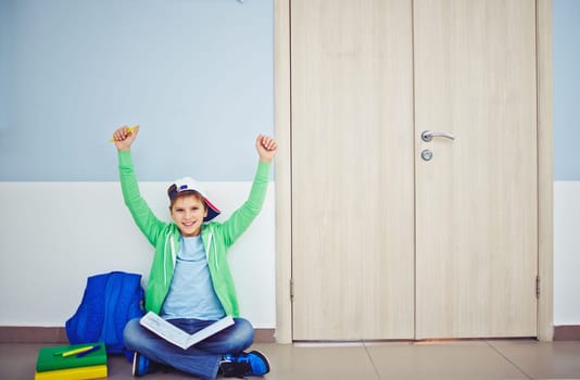 Ecstatic schoolboy with book and raised arms sitting on the floor by classroom door