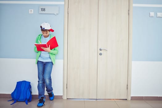 Cute schoolboy writing notes in exercise-book while standing by classroom door