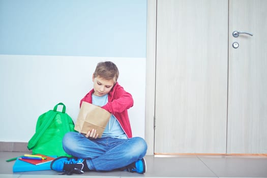 Cute schoolboy unpacking his lunch while sitting on the floor by classroom door