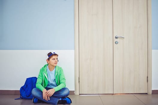 Cute schoolboy with backpack sitting on the floor by classroom door