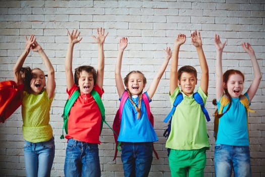 Several ecstatic classmates with backpacks raising arms against brick wall