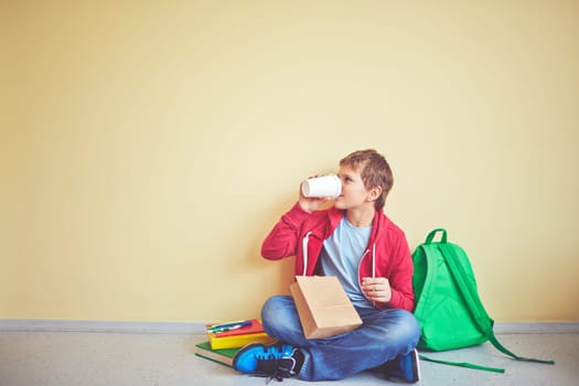 Handsome youngster sitting by the wall of school corridor and drinking