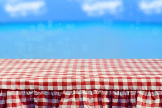 Empty wooden deck table with red checkered tablecloth over abstract bright blue sky and clouds background. For your food and product display montage.