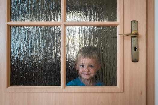 Portrait of a cute little Caucasian boy hiding behind a door with glass windows while playing hide and seek