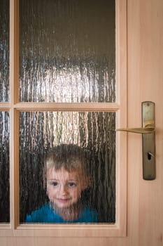 Portrait of a cute little Caucasian boy hiding behind a door with glass windows while playing hide and seek