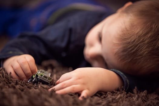 Cute little Caucasian boy playing with his tiny toy car while lying on the carpet in his room at home