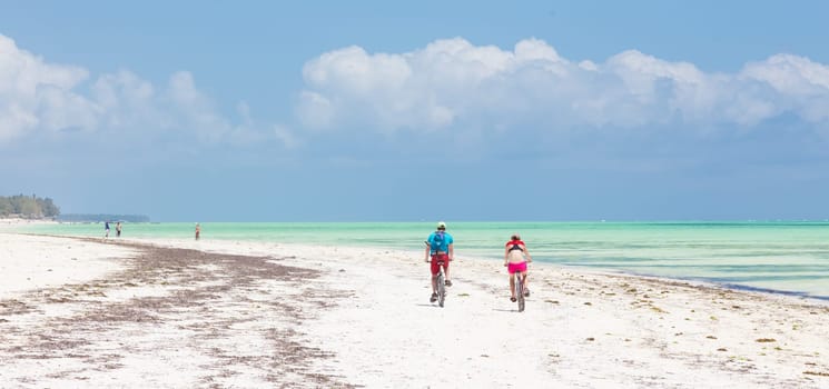 Active sporty tourist couple cycling down picture perfect white sand tropical beach of Paje village while being on vacations on Zanzibar, Tanzania.