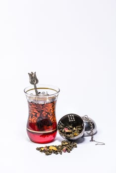 Typical Turkish glass for tea with a teaspoon and tea inside and an open infuserwith mango green tea on a white background