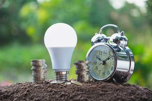 Energy saving light bulb and alarm clock on stacks of coins on nature background. Saving, accounting and financial concept.