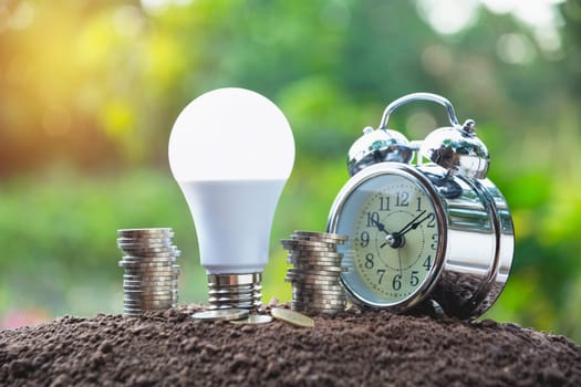 Energy saving light bulb and alarm clock on stacks of coins on nature background. Saving, accounting and financial concept.
